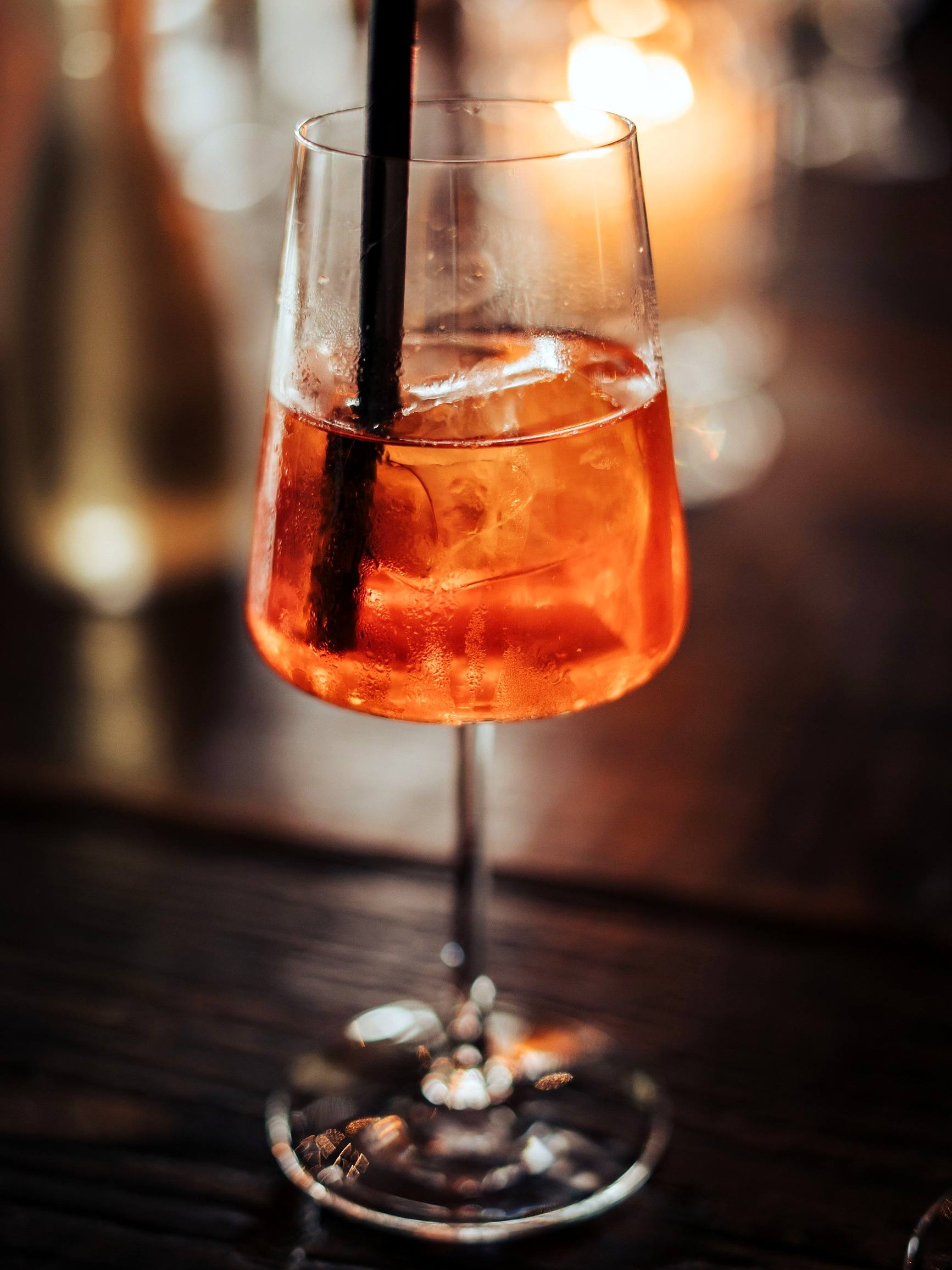 Aperol Spritz mit Eis und schwarzem Strohhalm im Stielglas, dahinter warmes Kerzenlicht